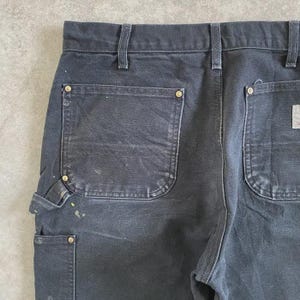 Puede incluir: Primer plano de la parte trasera de unos pantalones de trabajo negros. Los pantalones tienen bolsillos traseros grandes, un peque&ntilde;o bolsillo utilitario y remaches de lat&oacute;n. Salpicaduras de pintura a&ntilde;aden un aspecto desgastado a esta duradera ropa de trabajo.