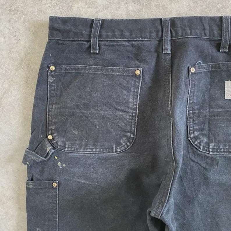 Puede incluir: Primer plano de la parte trasera de unos pantalones de trabajo negros. Los pantalones tienen bolsillos traseros grandes, un peque&ntilde;o bolsillo utilitario y remaches de lat&oacute;n. Salpicaduras de pintura a&ntilde;aden un aspecto desgastado a esta duradera ropa de trabajo.