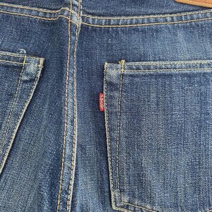 May include: Close-up of the back of blue denim jeans. The image shows the back pockets, stitching, and a small red tag. The denim has a classic, worn look.