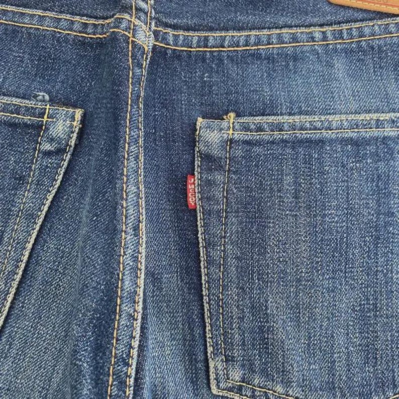 May include: Close-up of the back of blue denim jeans. The image shows the back pockets, stitching, and a small red tag. The denim has a classic, worn look.