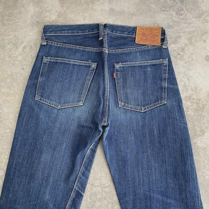 May include: Blue denim jeans with two back pockets and a small red tag. The jeans have a classic design with a brown leather patch on the back. The jeans are made of a durable denim material.