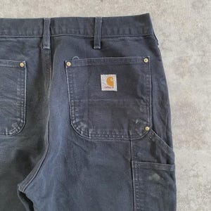 Puede incluir: Primer plano de un pantal&oacute;n de trabajo negro con el logotipo de Carhartt en el bolsillo trasero. El pantal&oacute;n tiene m&uacute;ltiples bolsillos y remaches de lat&oacute;n. La tela muestra signos de desgaste.