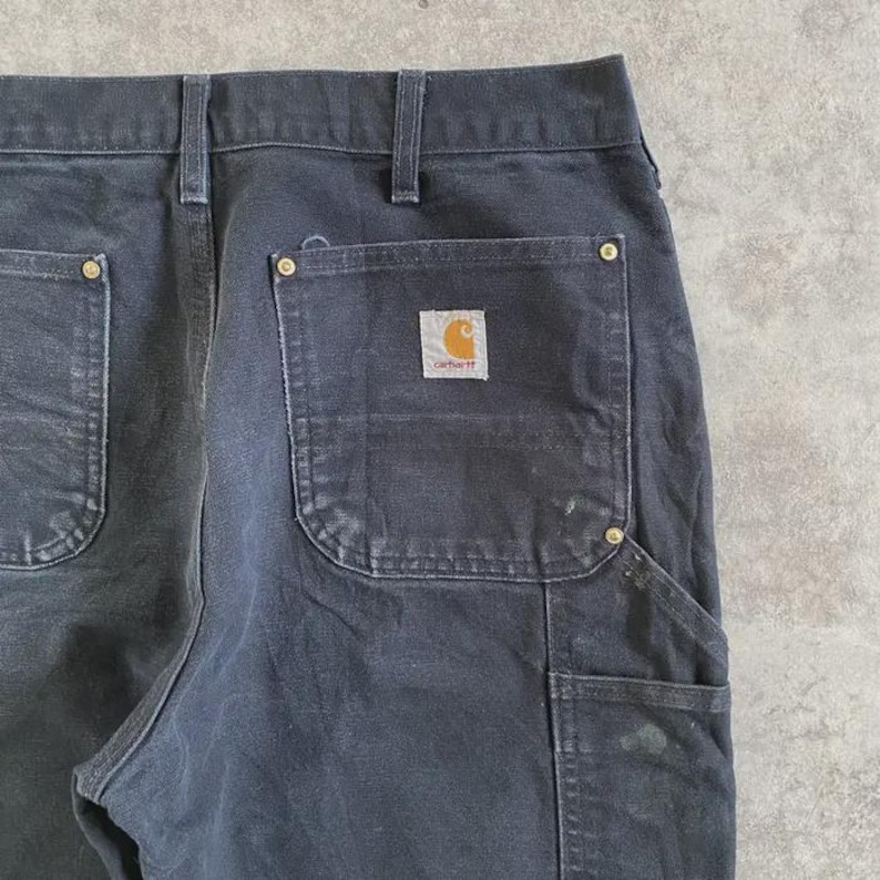Puede incluir: Primer plano de un pantal&oacute;n de trabajo negro con el logotipo de Carhartt en el bolsillo trasero. El pantal&oacute;n tiene m&uacute;ltiples bolsillos y remaches de lat&oacute;n. La tela muestra signos de desgaste.