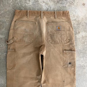 K&ouml;nnte beinhalten: Nahaufnahme einer beigefarbenen Arbeitshose mit mehreren Taschen und einem Carhartt-Logo-Patch. Die Hose weist Gebrauchsspuren auf, mit sichtbaren Flecken und Ausbleichen. Das Design umfasst eine Hammerschlaufe und verst&auml;rkte Taschen.