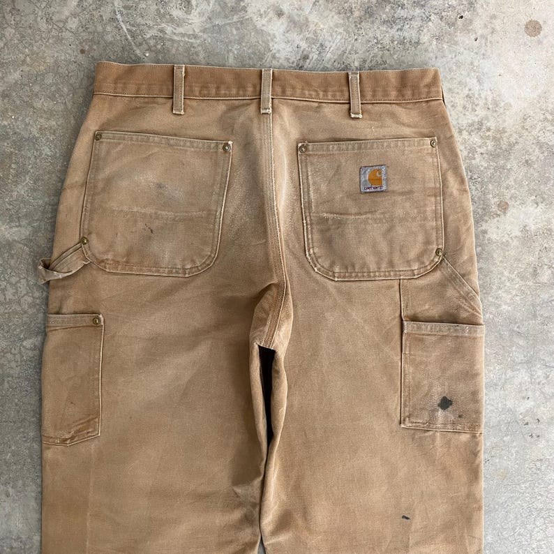 K&ouml;nnte beinhalten: Nahaufnahme einer beigefarbenen Arbeitshose mit mehreren Taschen und einem Carhartt-Logo-Patch. Die Hose weist Gebrauchsspuren auf, mit sichtbaren Flecken und Ausbleichen. Das Design umfasst eine Hammerschlaufe und verst&auml;rkte Taschen.