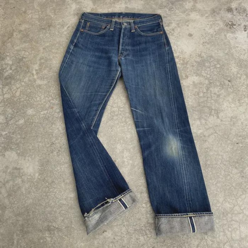 May include: Faded blue denim jeans with a classic five-pocket design. The jeans have a button fly, belt loops, and a straight leg fit. The cuffs are rolled up, revealing a contrasting gray and white fabric. The denim shows signs of wear.