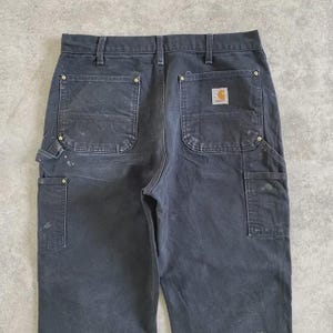 Puede incluir: Pantalones de trabajo gris oscuro con m&uacute;ltiples bolsillos y un parche con el logotipo de Carhartt en la parte trasera. Los pantalones muestran signos de desgaste, incluyendo salpicaduras de pintura. Incluyen trabilla para martillo y bolsillos reforzados.