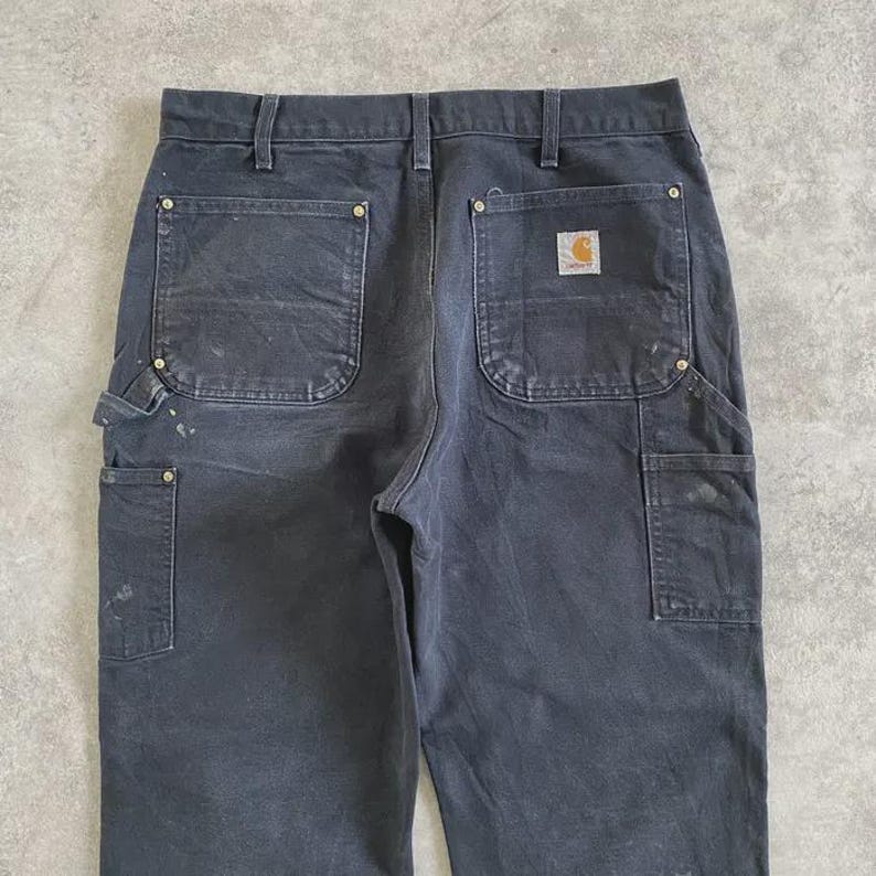 Puede incluir: Pantalones de trabajo gris oscuro con m&uacute;ltiples bolsillos y un parche con el logotipo de Carhartt en la parte trasera. Los pantalones muestran signos de desgaste, incluyendo salpicaduras de pintura. Incluyen trabilla para martillo y bolsillos reforzados.