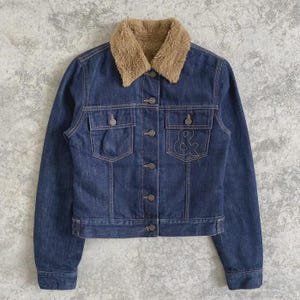 May include: A blue denim jacket with a tan faux fur collar. The jacket has two chest pockets, two side pockets, and a button-up closure. An embroidered ampersand is on the right pocket.