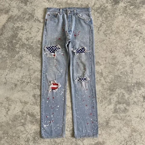May include: Light blue denim jeans with distressed patches featuring American flag designs. The jeans have a button fly and are splattered with red and white paint. A casual, patriotic style.