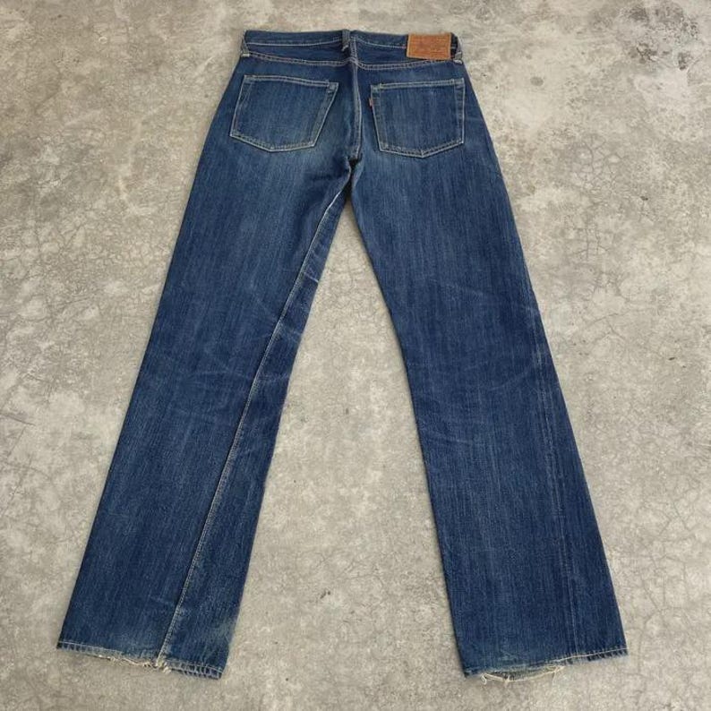 May include: A pair of blue denim jeans, viewed from the back, with two back pockets and a small leather patch. The jeans have a classic five-pocket design and a straight leg fit. The denim shows signs of wear and fading.