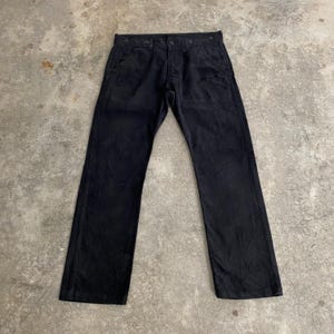 May include: Black pants with a button fly and multiple pockets. The pants are made of a textured fabric and have a straight leg design. The pants are laid flat on a concrete surface.