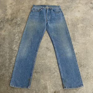 Könnte beinhalten: Eine mittelblaue Jeans aus Denim mit klassischem Fünf-Taschen-Design, Knopfleiste, Gürtelschlaufen am Bund und gerade geschnittenen Beinen mit subtilen Verwaschungen, die dem Stoff ein getragenes Aussehen verleihen.