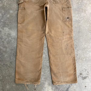 K&ouml;nnte beinhalten: Beige Arbeitshose mit mehreren Taschen und einem abgenutzten Aussehen. Die Hose weist Gebrauchsspuren auf, darunter Flecken und ausgefranste Kanten. Die Arbeitshose ist aus einem strapazierf&auml;higen Material gefertigt.