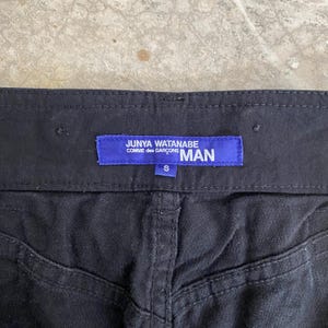 May include: Close-up of black pants with a visible waistband and stitching details. A blue label reads "JUNYA WATANABE COMME des GAR&Ccedil;ONS MAN" with a size "S" tag.