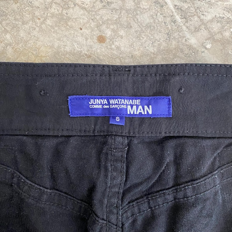 May include: Close-up of black pants with a visible waistband and stitching details. A blue label reads "JUNYA WATANABE COMME des GAR&Ccedil;ONS MAN" with a size "S" tag.