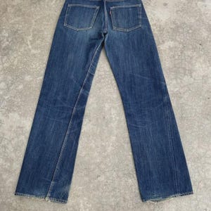 May include: A pair of vintage blue denim jeans, viewed from the back. The jeans feature two back pockets, a red tag, and a classic straight leg fit. The denim shows signs of wear and fading, adding to their vintage appeal.