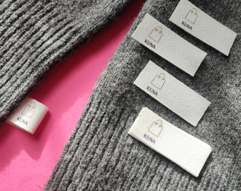 May include: Four white fabric labels with the word "KUNA" and a small shopping bag icon printed in brown. The labels are attached to a grey knitted fabric.