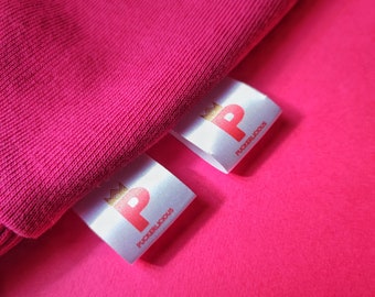 May include: Two white fabric labels with a pink "P" and the text "Puckerlicious" in gold lettering. The labels are on a pink fabric surface.