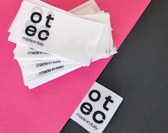 May include: A pile of white fabric labels, each bearing the text "ot ec made in Italy" in black. The labels are arranged on a pink and black surface.