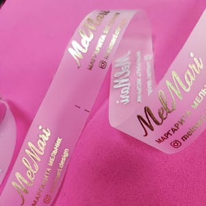 May include: A pink ribbon with gold lettering that reads "Mel Mari" and other text. The ribbon is curled and lies on a pink surface. The text includes a name, a social media handle, and a website address.