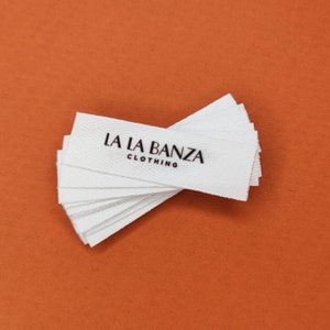 May include: A stack of white woven clothing labels with the text "LA LA BANZA CLOTHING" printed in black.