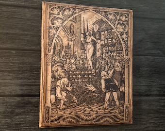 May include: A rectangular wooden plaque with a detailed engraving depicting a medieval scene. The artwork features figures in period clothing, buildings, and an archway. The plaque has a decorative border and is set against a dark wood background.
