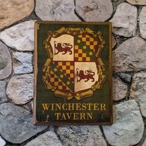 May include: A wooden sign with a green background and a gold and red crest featuring two lions. The sign reads "Winchester Tavern".