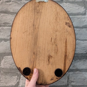 May include: The back of a wooden oval sign with a metal hanger and black felt pads on the bottom.
