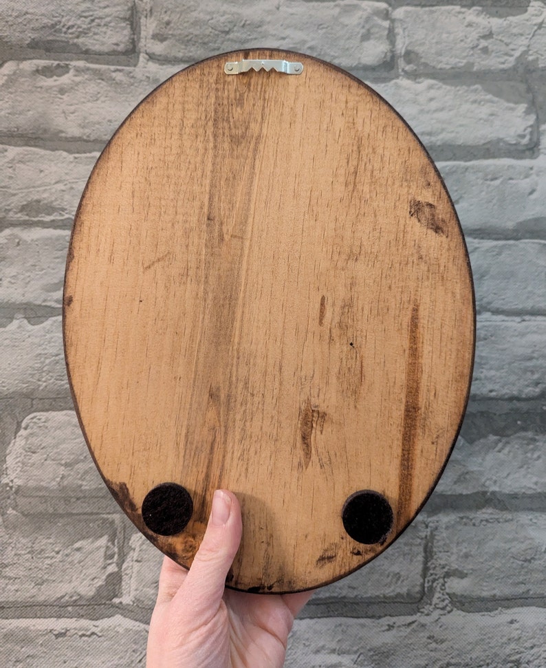 May include: The back of a wooden oval sign with a metal hanger and black felt pads on the bottom.