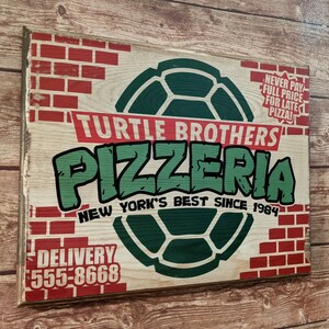 May include: Wooden sign for Turtle Brothers Pizzeria, featuring a green turtle shell design and the words "Pizzeria" and "New York's Best Since 1984." The sign has a distressed look with red brick accents and the phone number 555-8668.