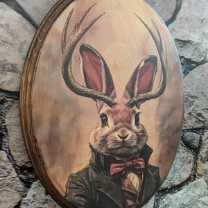 May include: Oval wooden plaque featuring a rabbit with deer antlers, dressed in a black coat and red bow tie. The rabbit has large ears and is set against a sepia-toned background. The plaque has a dark brown wooden frame.