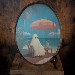 May include: Oval wooden art piece featuring a ghost relaxing on a beach. The ghost is seated in a beach chair under a coral-colored umbrella, with a drink on a small table. The background shows a blue ocean and sky.