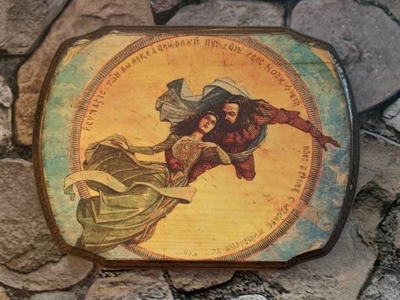 May include: A circular wooden plaque with a painted image of a woman and a man flying through the air. The woman is wearing a long flowing dress and the man is wearing a dark robe. The plaque has a blue and white border with text in a foreign language.