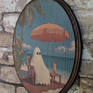 May include: Round wooden wall art featuring a ghost relaxing on a beach under an orange umbrella. The ghost is sitting in a wooden chair, with a drink on a small table. The background shows a blue ocean and palm trees.