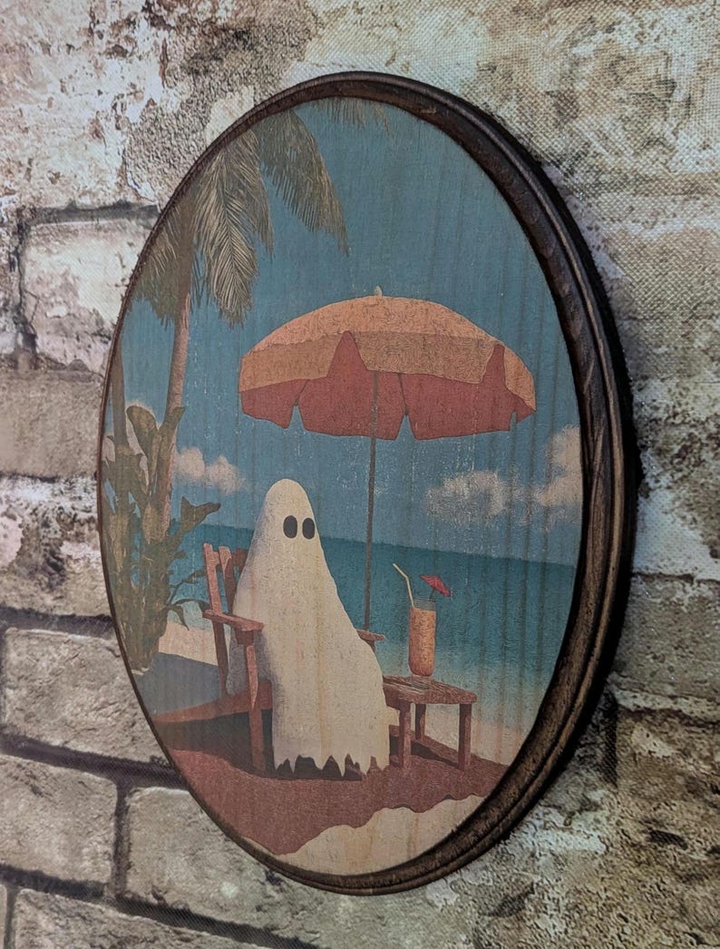 May include: Round wooden wall art featuring a ghost relaxing on a beach under an orange umbrella. The ghost is sitting in a wooden chair, with a drink on a small table. The background shows a blue ocean and palm trees.