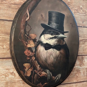 K&ouml;nnte beinhalten: Eine runde Holzplakette mit einem Gem&auml;lde eines Vogels, der einen schwarzen Zylinder und eine Fliege tr&auml;gt. Der Vogel sitzt auf einem Ast mit braunen und gelben Bl&auml;ttern. Der Hintergrund ist dunkelbraun.