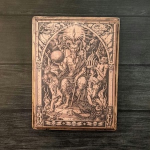 Puede incluir: Una ilustración en blanco y negro de una figura con cuernos sentada en un trono rodeada de otras figuras. La figura sostiene una esfera en una mano y un bastón en la otra. La ilustración está enmarcada en un marco de madera.