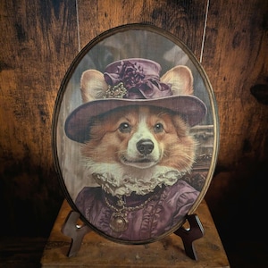 May include: An oval portrait of a Corgi dog in a purple hat and dress, styled like a Victorian painting. The dog has a dignified expression, with detailed fur and clothing. The portrait is set in a wooden frame, displayed on a wooden stand.