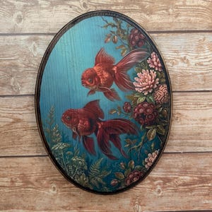 May include: Oval wooden wall art featuring two red goldfish swimming in a blue aquatic scene. The artwork is framed with a dark brown border and includes floral and plant details in shades of pink, red, and green.