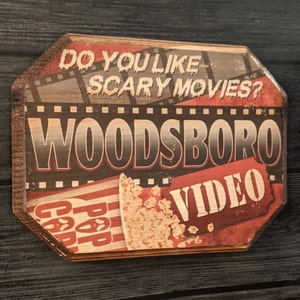 May include: A vintage-style wooden sign with the text "Do you like scary movies? Woodsboro Video" in red and black lettering. The sign features a red and white striped popcorn bucket with popcorn spilling out.