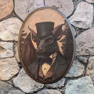May include: A black bat wearing a top hat and a bow tie, with a brown and gold patterned vest, in a framed oval portrait.