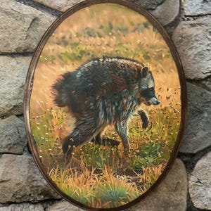 May include: An oval-shaped wooden plaque featuring a detailed painting of a raccoon walking through tall, golden-brown grasses. The raccoon has dark fur with characteristic facial markings, and the artwork is framed by a smooth, dark wood border.