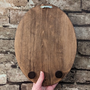 May include: Back of a stained, oval wooden plaque with a metal sawtooth hanger. Two small, round, felt pads are attached to the bottom. The wood grain is visible. The plaque is held by a hand.
