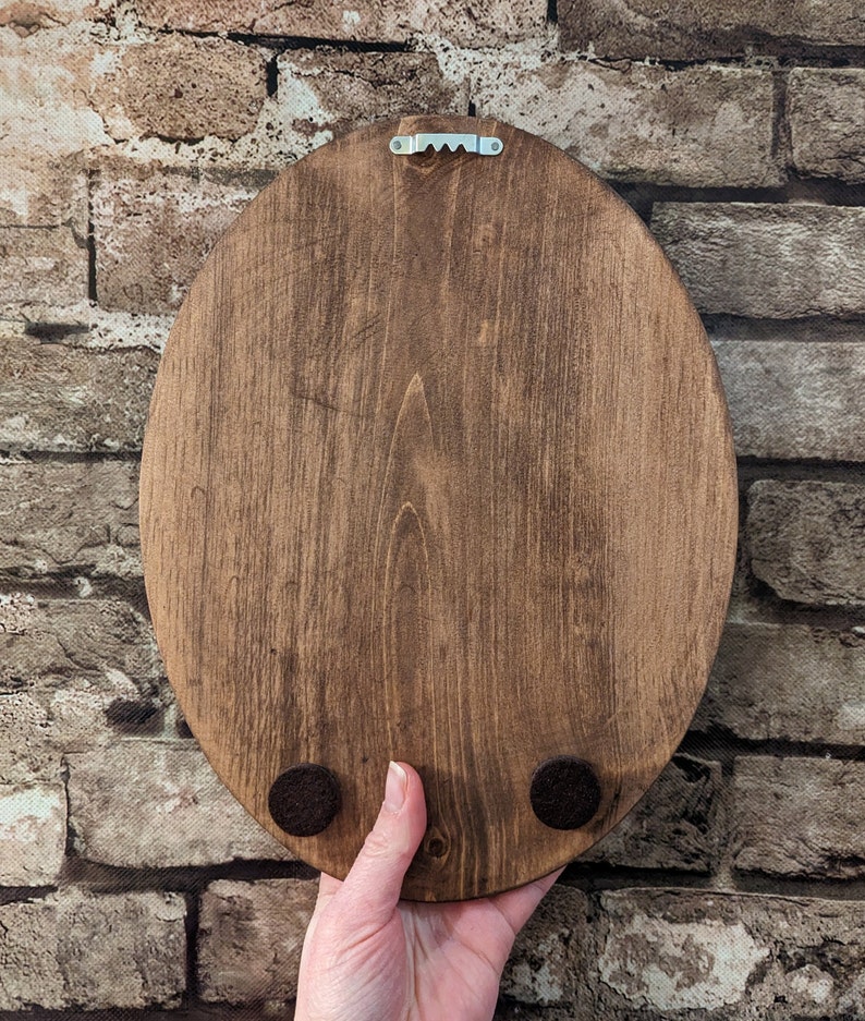 May include: Back of a stained, oval wooden plaque with a metal sawtooth hanger. Two small, round, felt pads are attached to the bottom. The wood grain is visible. The plaque is held by a hand.