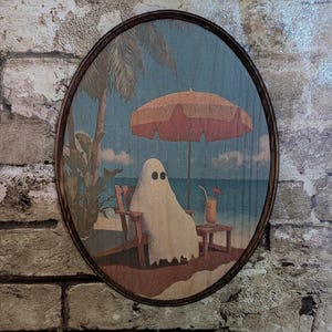 May include: Oval wooden wall art featuring a ghost relaxing on a beach under a coral umbrella. The ghost is seated in a wooden beach chair, with a drink on a small table. Palm trees and ocean in the background.