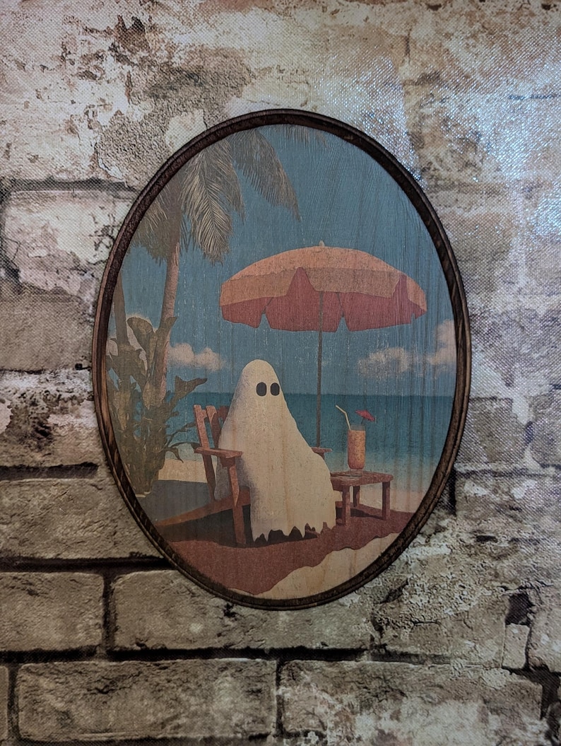 May include: Oval wooden wall art featuring a ghost relaxing on a beach under a coral umbrella. The ghost is seated in a wooden beach chair, with a drink on a small table. Palm trees and ocean in the background.