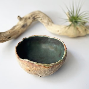Può includere: Una piccola ciotola in ceramica rustica con una glassa verde scuro e un esterno ruvido e testurizzato. La ciotola è su una superficie bianca.