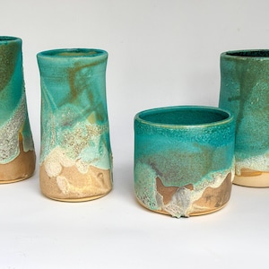 Op de afbeelding: Vier handgemaakte keramische vazen en bekers in verschillende vormen. Het aardewerk heeft een turquoise en groene glazuur met een zanderige, getextureerde basis. De vaten hebben een unieke, organische esthetiek, perfect voor de interieurdecoratie.