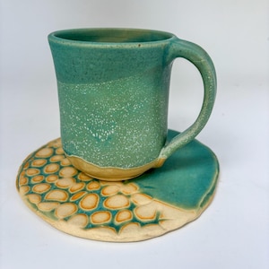 Peut inclure: Un mug en céramique et son dessous de plat assorti. Le mug est de couleur turquoise avec une finition mouchetée et une anse incurvée. Le dessous de plat a un dessus turquoise et une base beige avec un motif orange et beige. Article de cuisine artisanal unique.